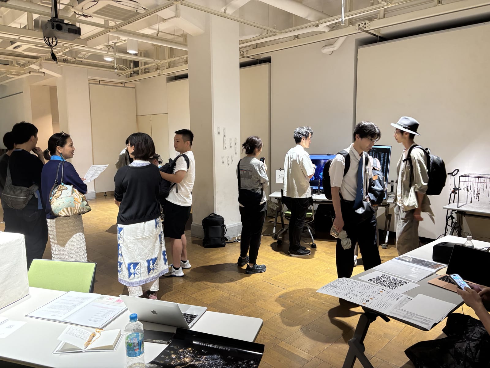 現代美術×ブロックチェーンSG成果発表会の会場風景。複数の出品作品の前に鑑賞者が集まり、中央奥のディスプレイに《めぐる種》が映っている